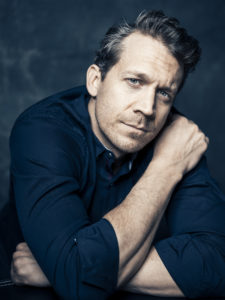 A stubble bearded white male reaching into his 40’s wearing a dark blue button shirt, sitting with his arms folded upright as if he is hugging himself. He stares directly into the lens of the camera into your soul. The backdrop is a cloudy blue.