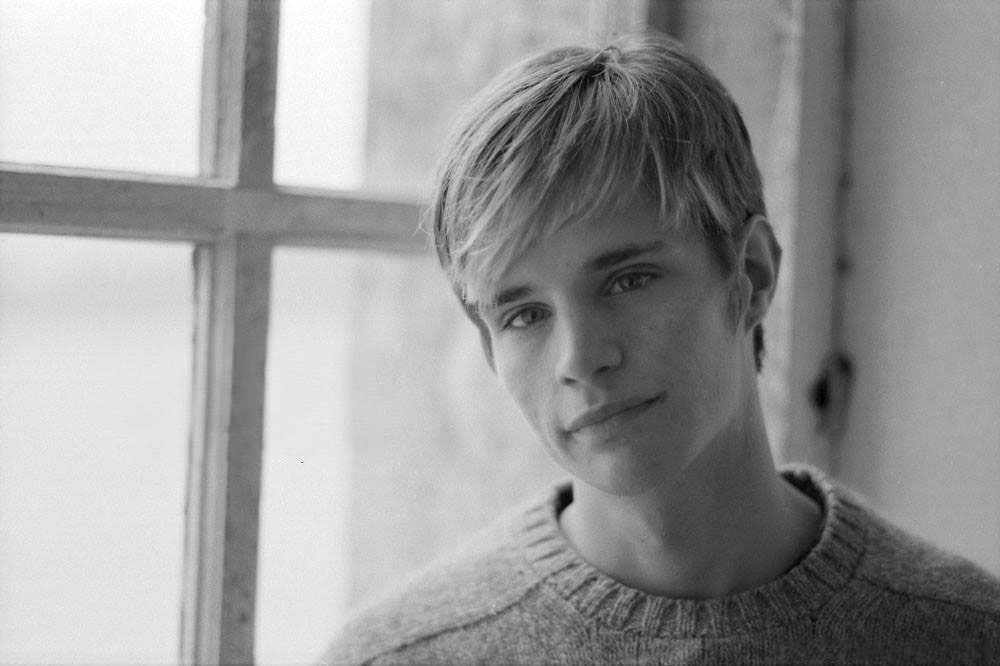 black and white photo of a young white male in his early 20s. he has blond hair that is combed over. He wears a sweater and stands in front of a large window.